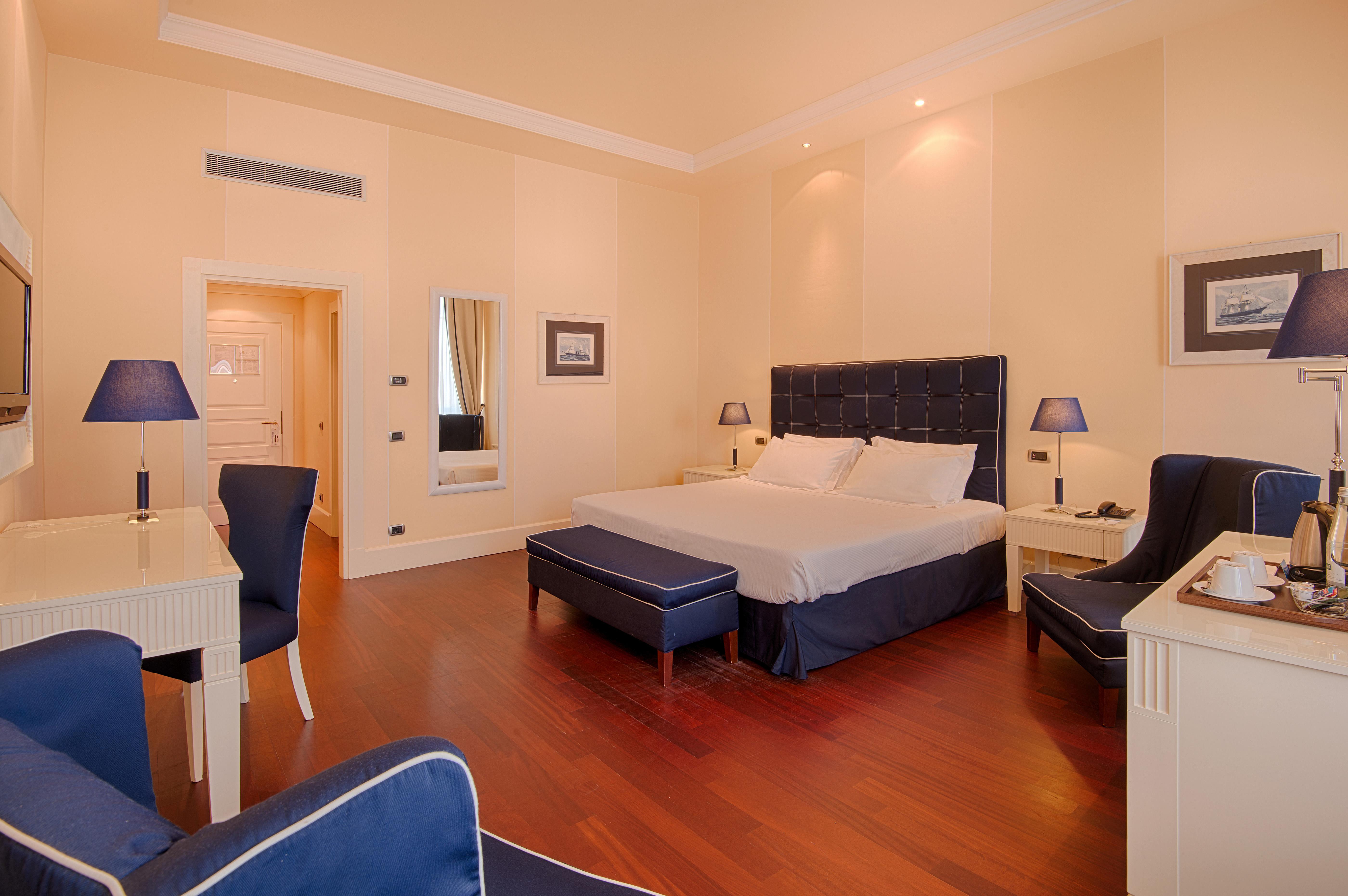 Gh Palazzo Suite & Spa Λιβόρνο Εξωτερικό φωτογραφία The photo shows a stylish hotel room. It features a large bed with white linens and dark blue upholstered headboard. On either side of the bed, there are bedside tables with lamps. The room has wooden flooring and light-colored walls, creating a brig