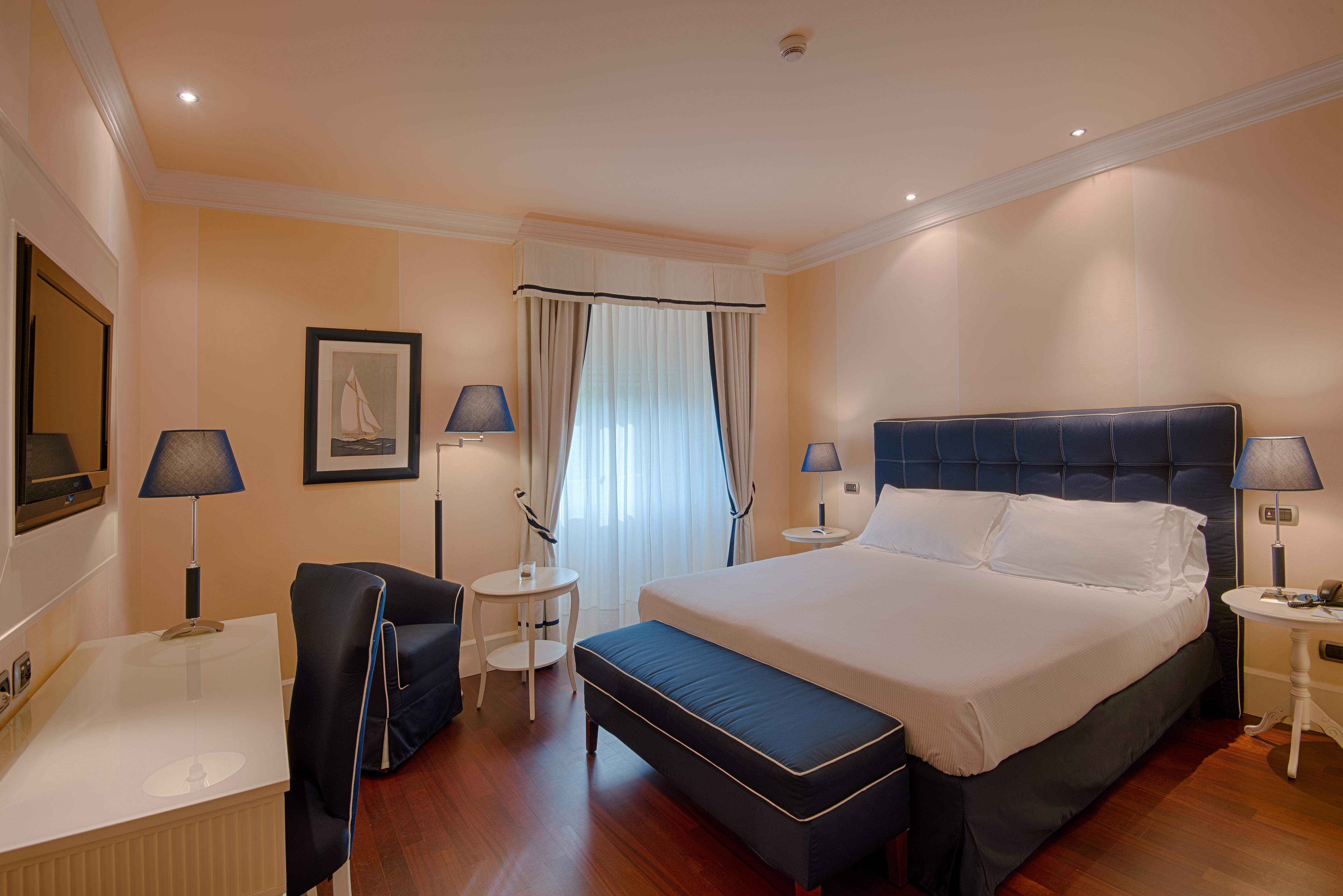 Gh Palazzo Suite & Spa Λιβόρνο Εξωτερικό φωτογραφία The photo shows a well-furnished hotel room. It features a large bed with a blue headboard, white bedding, and a matching blue ottoman at the foot of the bed. There are two bedside lamps on small tables next to the bed. A comfortable armchair is posi
