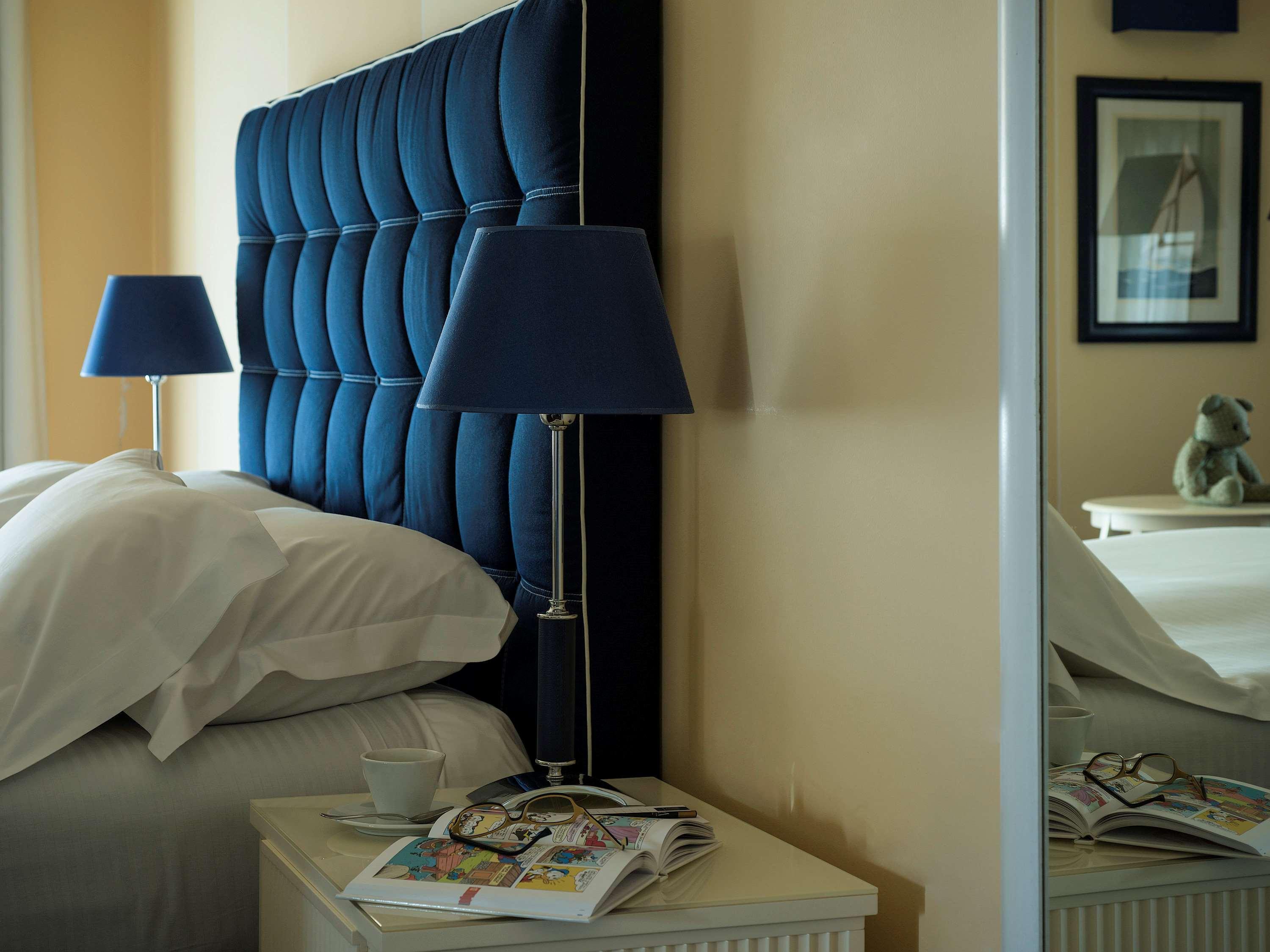 Gh Palazzo Suite & Spa Λιβόρνο Εξωτερικό φωτογραφία The photo shows a cozy bedroom setting. There is a large, tufted headboard in a deep blue color behind a neatly made bed with white bedding. On either side of the bed are bedside tables, each featuring a blue lamp. One table has a small cup and a sta