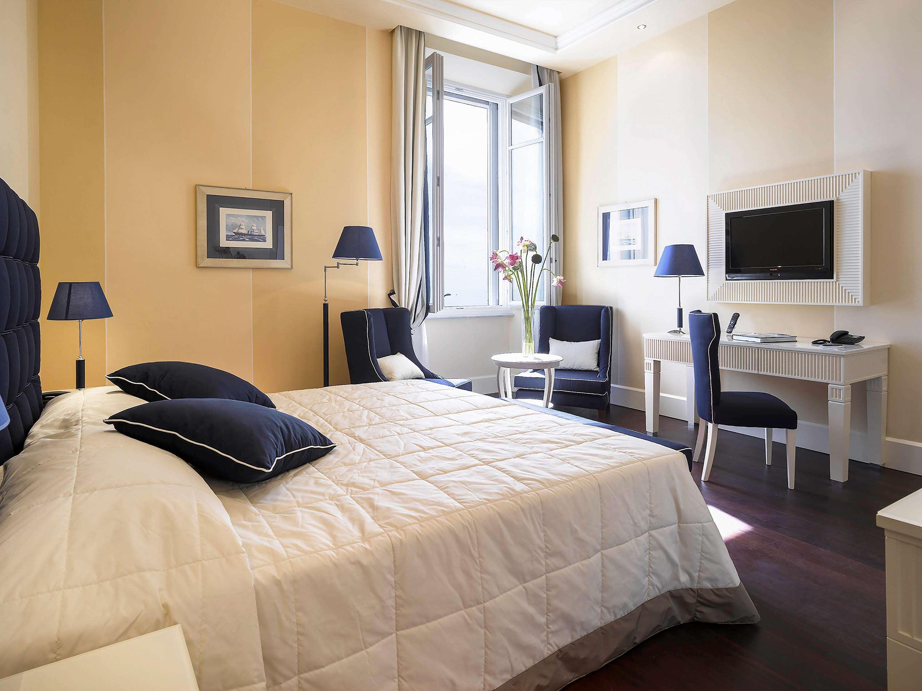 Gh Palazzo Suite & Spa Λιβόρνο Εξωτερικό φωτογραφία The photo shows a well-furnished hotel room. It features a large bed with a white quilt and blue accented pillows. The walls are painted a soft yellow, and there's a window that allows natural light to enter the space. Two blue chairs are positioned 