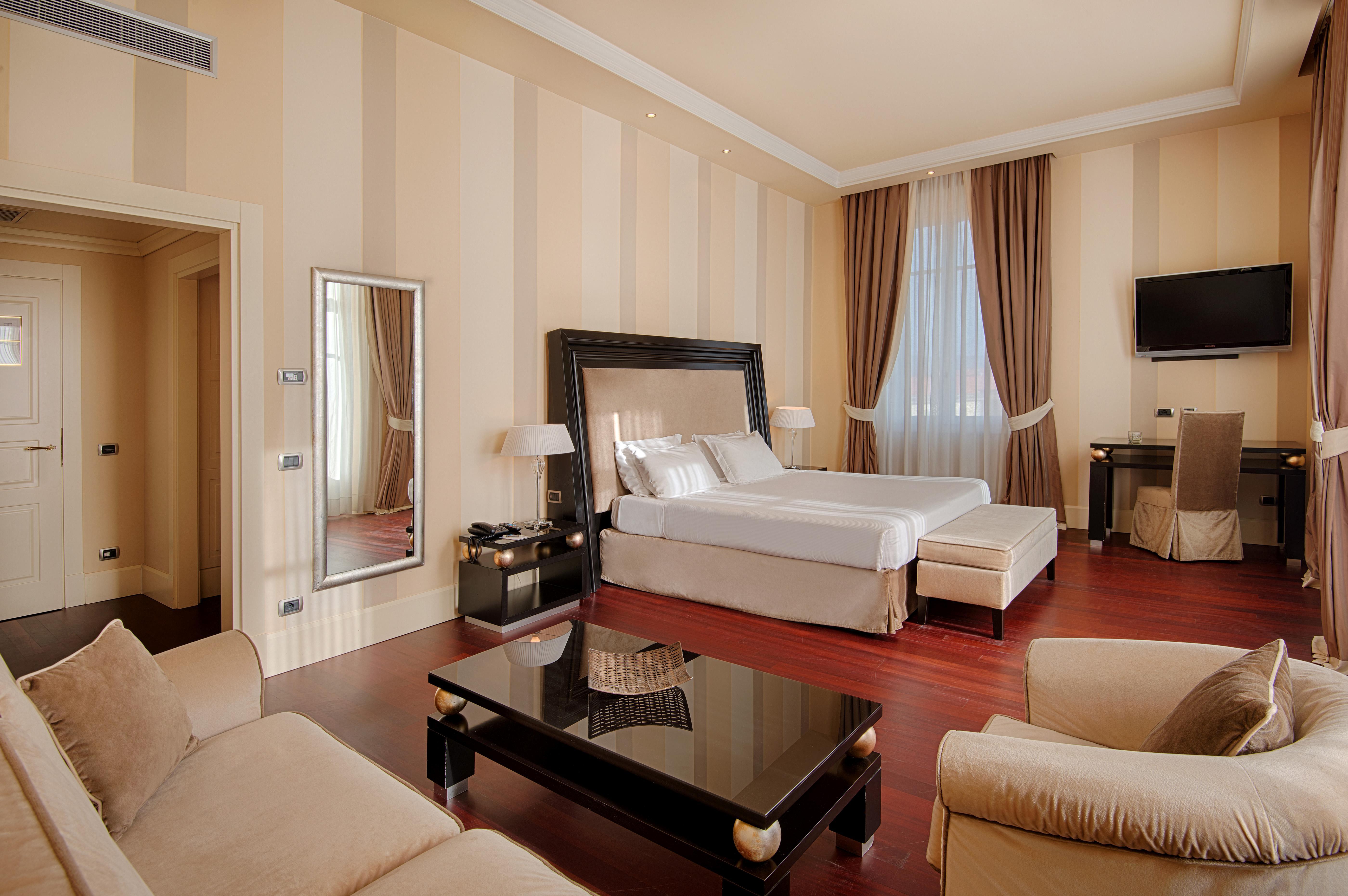 Gh Palazzo Suite & Spa Λιβόρνο Εξωτερικό φωτογραφία The photo shows a well-appointed hotel room. It features a large bed with crisp white linens, placed against a stylish wooden headboard. To the left, there is a full-length mirror and a small seating area with a cream-colored sofa and an armchair. Th