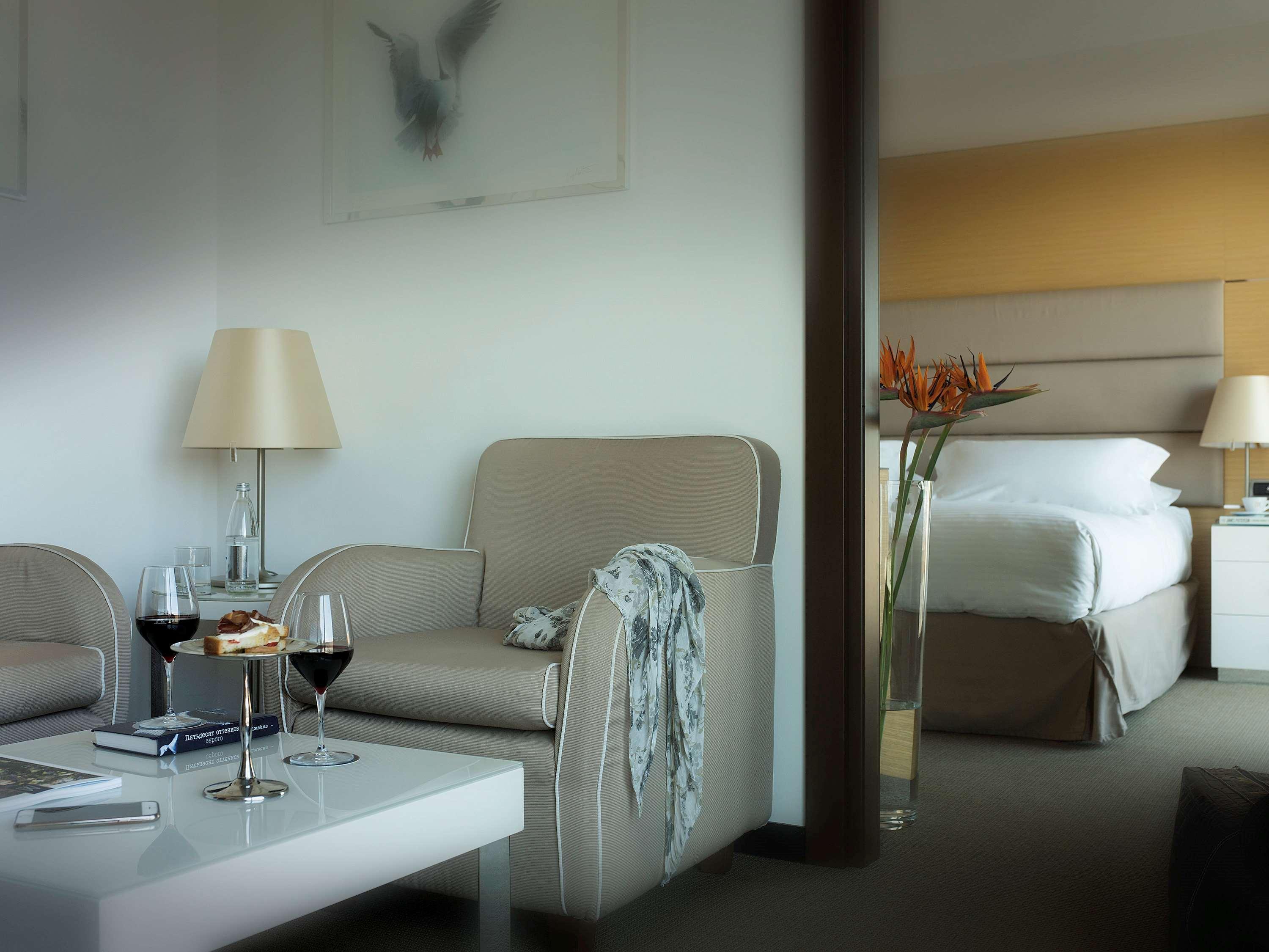 Gh Palazzo Suite & Spa Λιβόρνο Εξωτερικό φωτογραφία The photo shows a modern hotel room or suite. On one side, there is a cozy seating area with two light-colored chairs and a small coffee table. On the table, there are two glasses of red wine and a plate of cookies, suggesting a relaxing atmosphere. 