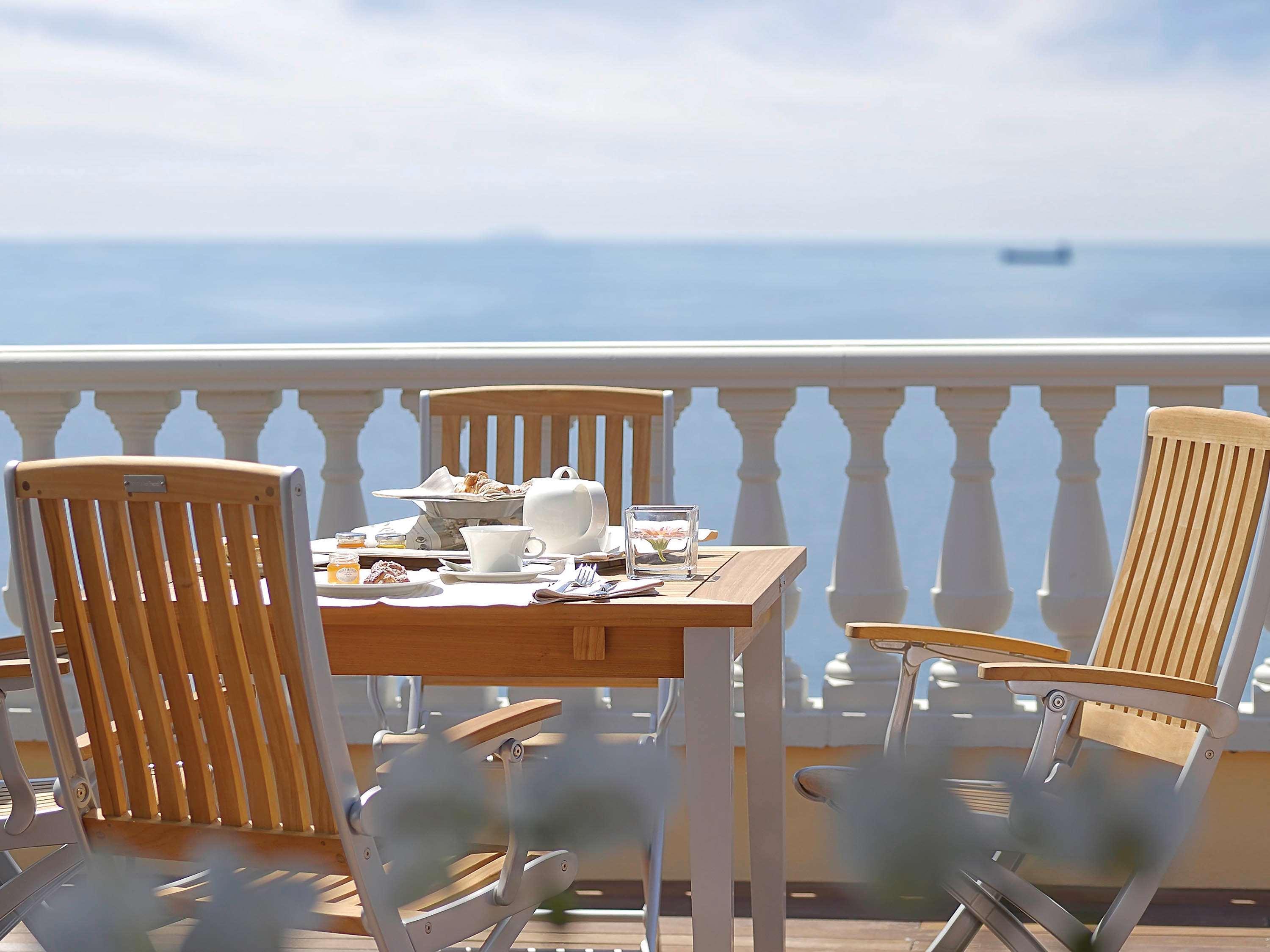 Gh Palazzo Suite & Spa Λιβόρνο Εξωτερικό φωτογραφία The photo shows a beautiful outdoor setting, likely a balcony or terrace overlooking a calm sea. There is a wooden table with a light breakfast spread, including a tea set and some food items. Two comfortable wooden chairs are positioned around the t